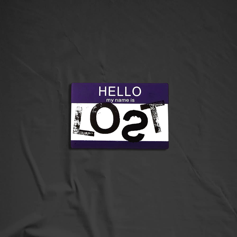 Hello my name is LOST sticker. A purple and white nametag sticker stamped with the name LOST with a backwards S. The sticker is set against a black background.