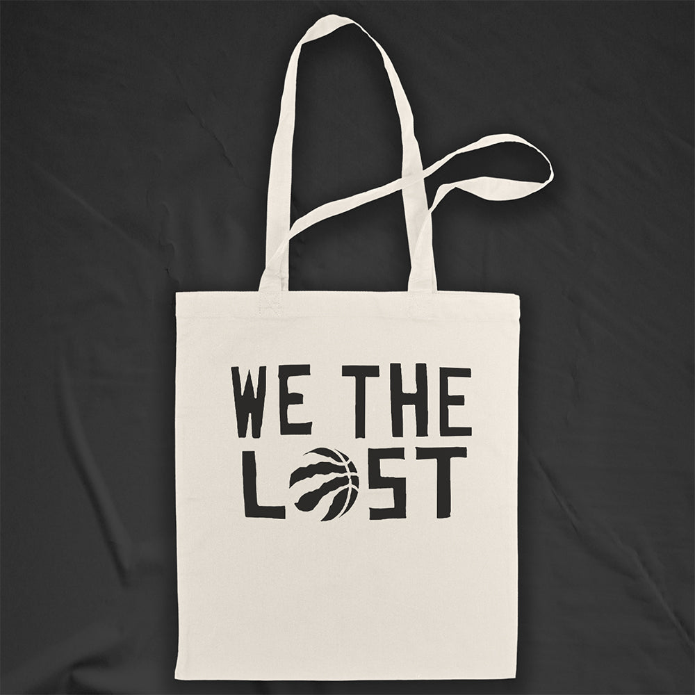 We the LOST printed in bold black lettering with the raptor claw design displayed on a natural canvas tote bag. A black background provides dramatic contrast to the tote bag on display.  