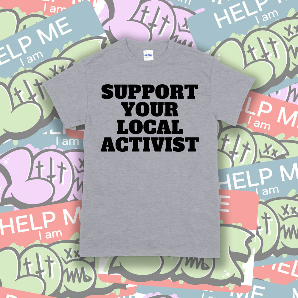 The original Support Your Local Activist t-shirt. Bold black letters screen printed on a gray shirt. The shirt is displayed against a wall of colorful graffiti stickers.