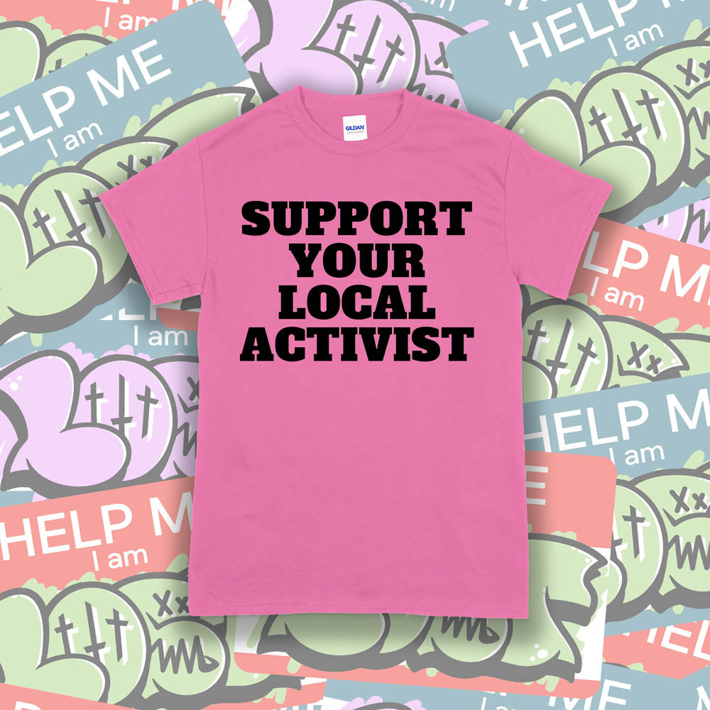 The original Support Your Local Activist t-shirt. Bold black letters screen printed on a pink shirt. The shirt is displayed against a wall of colorful graffiti stickers.